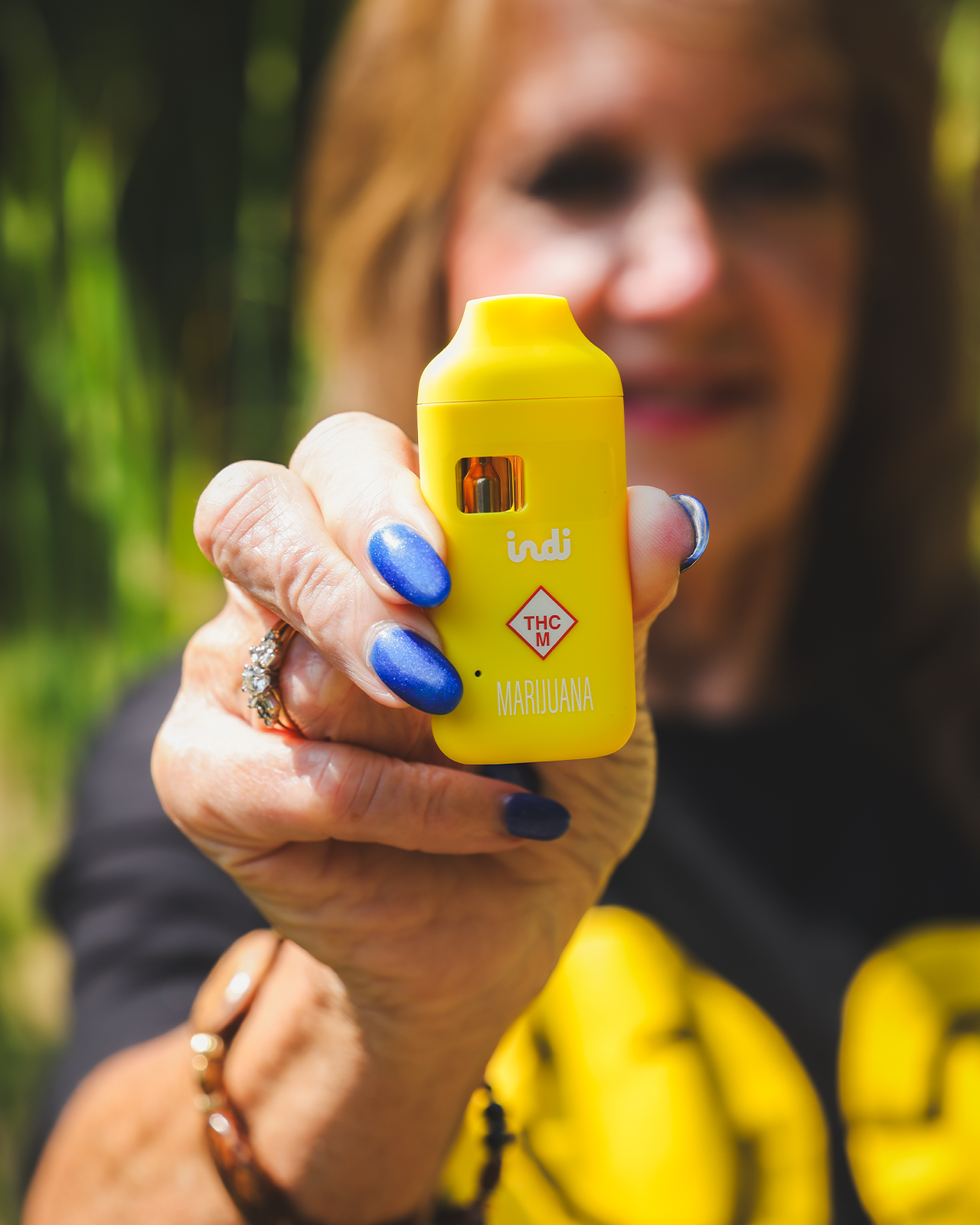 An older woman holds a yellow Indi vape and smiles in the background.