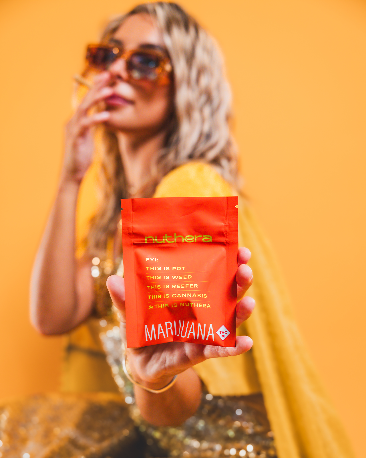 A woman with blonde hair wearing orange glasses holds a joint to her mouth while holding out Nuthera Flower's orange packaging.