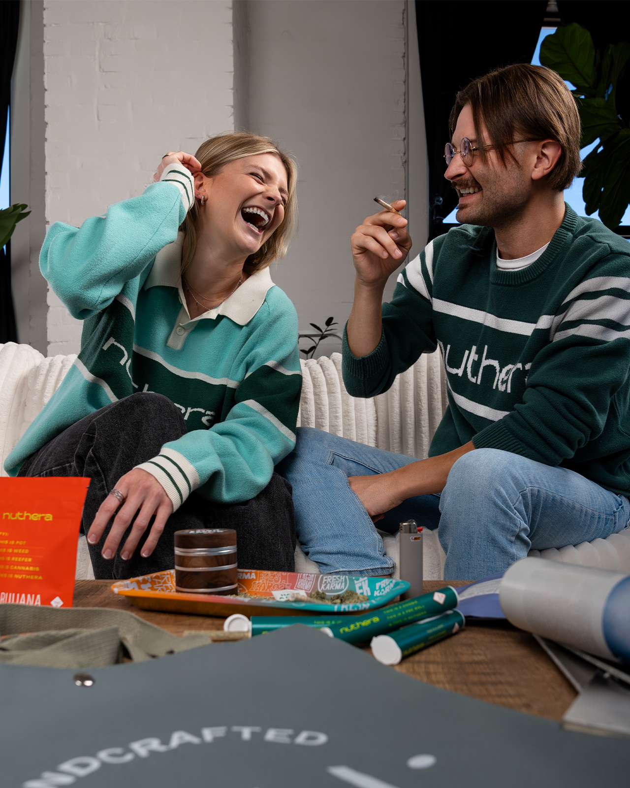 A woman and a man laugh while smoking a joint together. On the table is a rolling tray, grinder and sativa Nuthera packaging.
