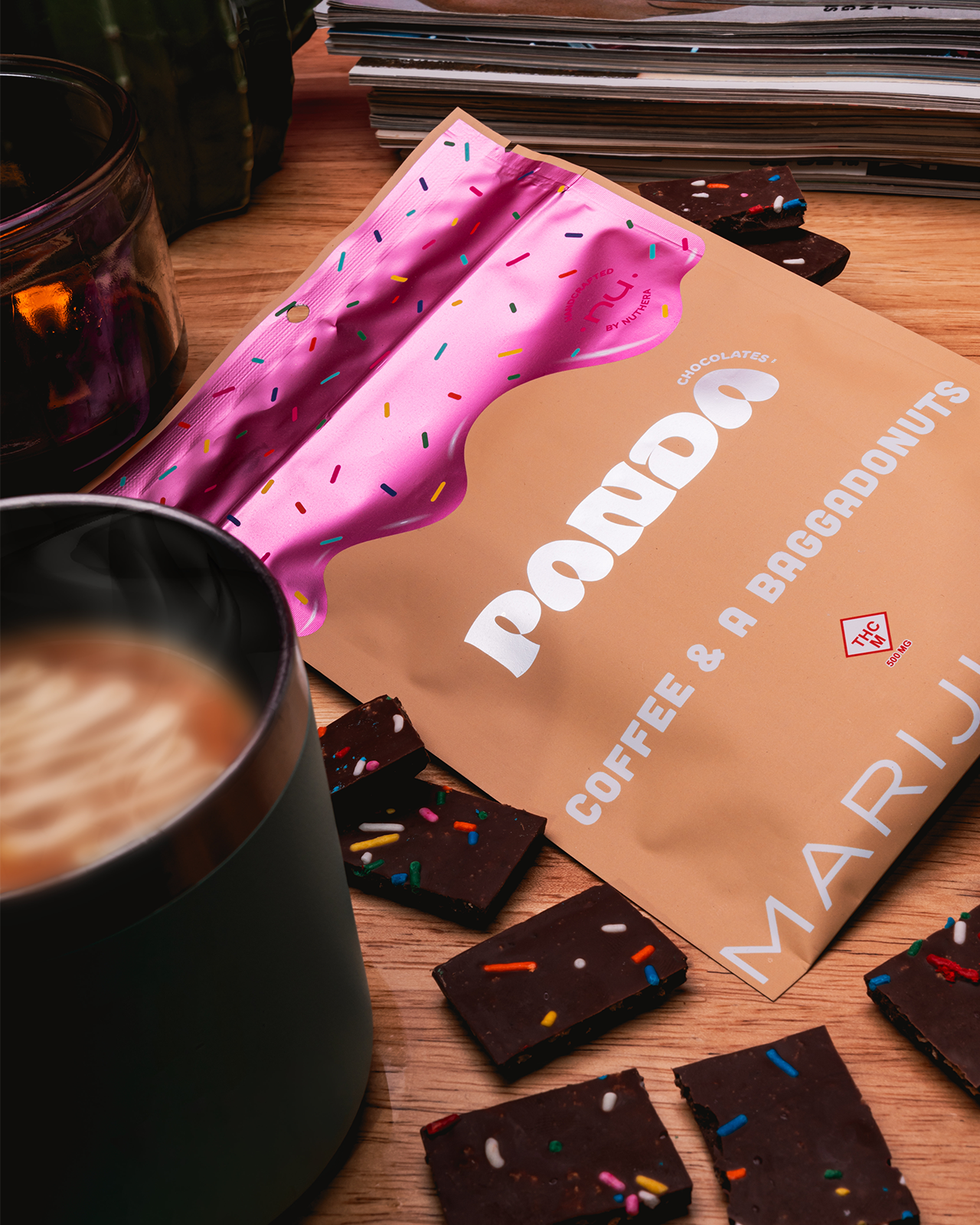 A package of the coffee & baggadonuts bar sits on a table next to a cup of coffee and the pieces of dark chocolate with sprinkles sit around the packaging.